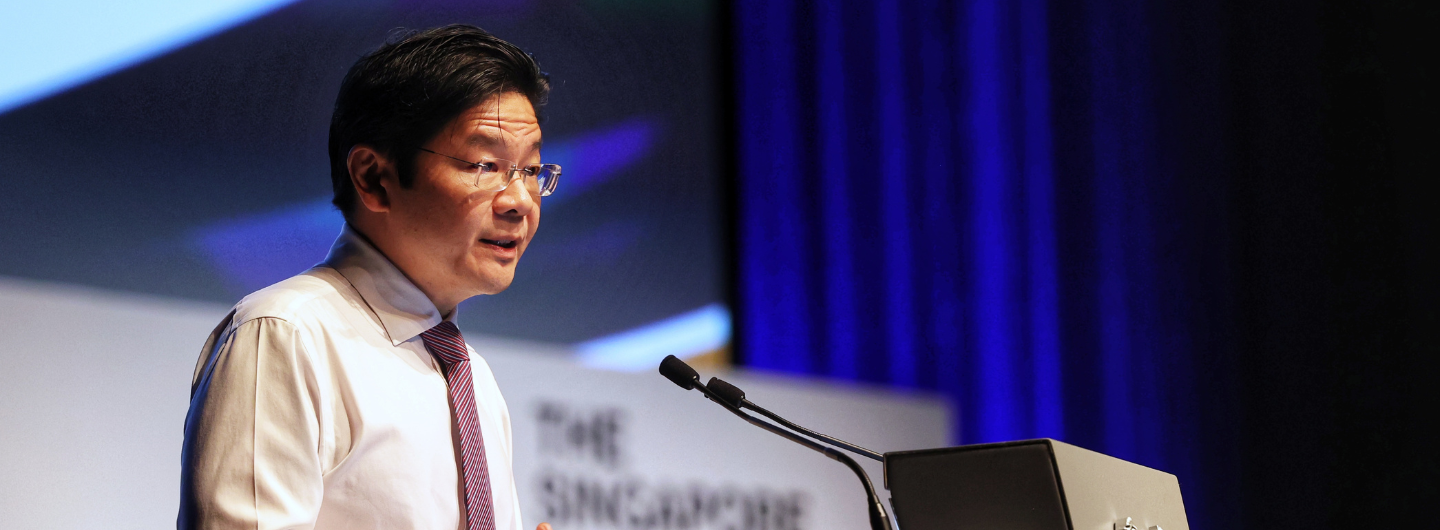 Singapore's Lawrence Wong at lectern, speaking into microphones, with blue draped background.